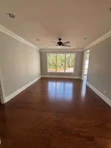 Bright living room with hardwood floors, light gray walls, white crown molding, recessed lighting, and a ceiling fan centered beneath a large window overlooking the outdoors.
