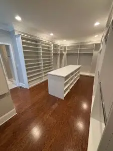 Spacious walk-in closet with hardwood floors, built-in white shelving and hanging racks along the walls, and a large central island with open storage beneath recessed lighting.