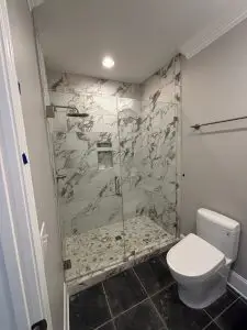 Elegant bathroom featuring a walk-in shower with marble-patterned tile walls, glass door enclosure, hexagon tile floor, and a modern white toilet on dark slate tile flooring.