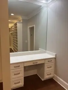 Built-in vanity area with white cabinetry, marble-style countertop, and large wall mirror leading into a spacious walk-in closet with custom shelving and hardwood floors.
